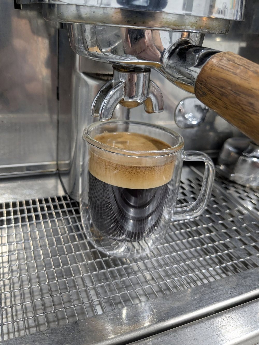 A double shot of espresso pulling into a clear glass mug, thick crema on top, under the chrome group head of the machine