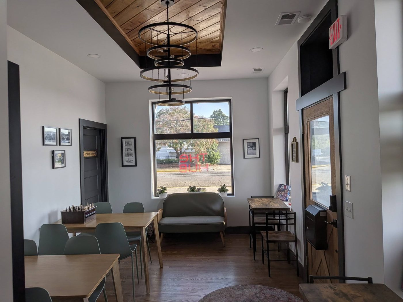 The Hub, the back room: a chandelier made of stacked metal rings under a wood ceiling, a green settee beneath the front window, a chess set on the table, and a neon sign reading The Hub in the glass