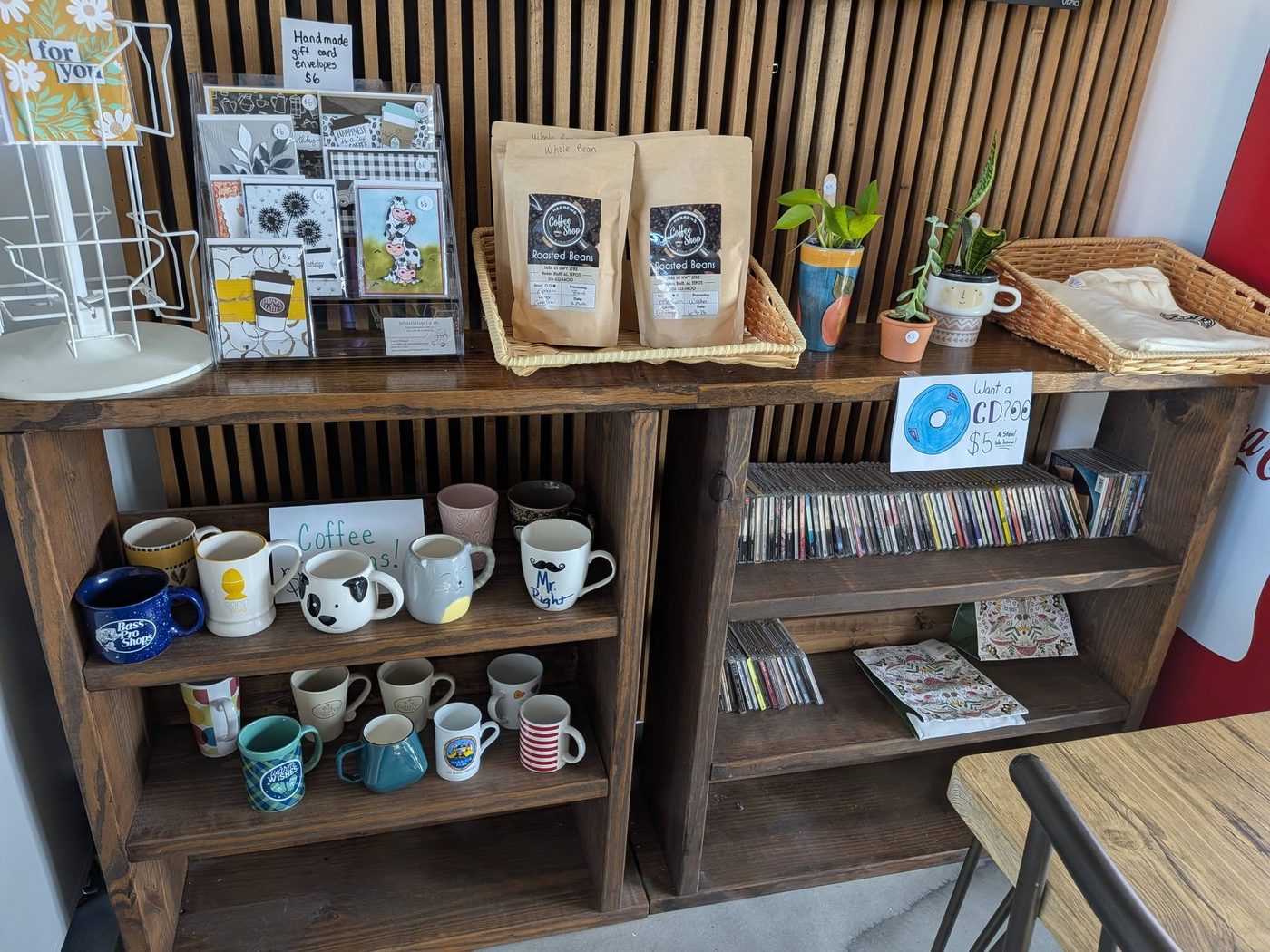 The retail shelf: a rack of handmade greeting cards, two bags of Hebrews Coffee Shop whole bean coffee in a basket, potted plants, a row of mugs, and a shelf of CDs with a hand-drawn sign
